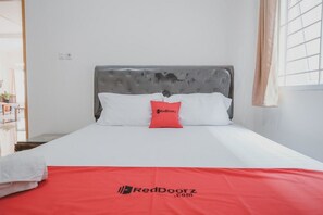 A neatly made bed with a grey upholstered headboard, white bedding, and a red pillow with the RedDoorz logo.