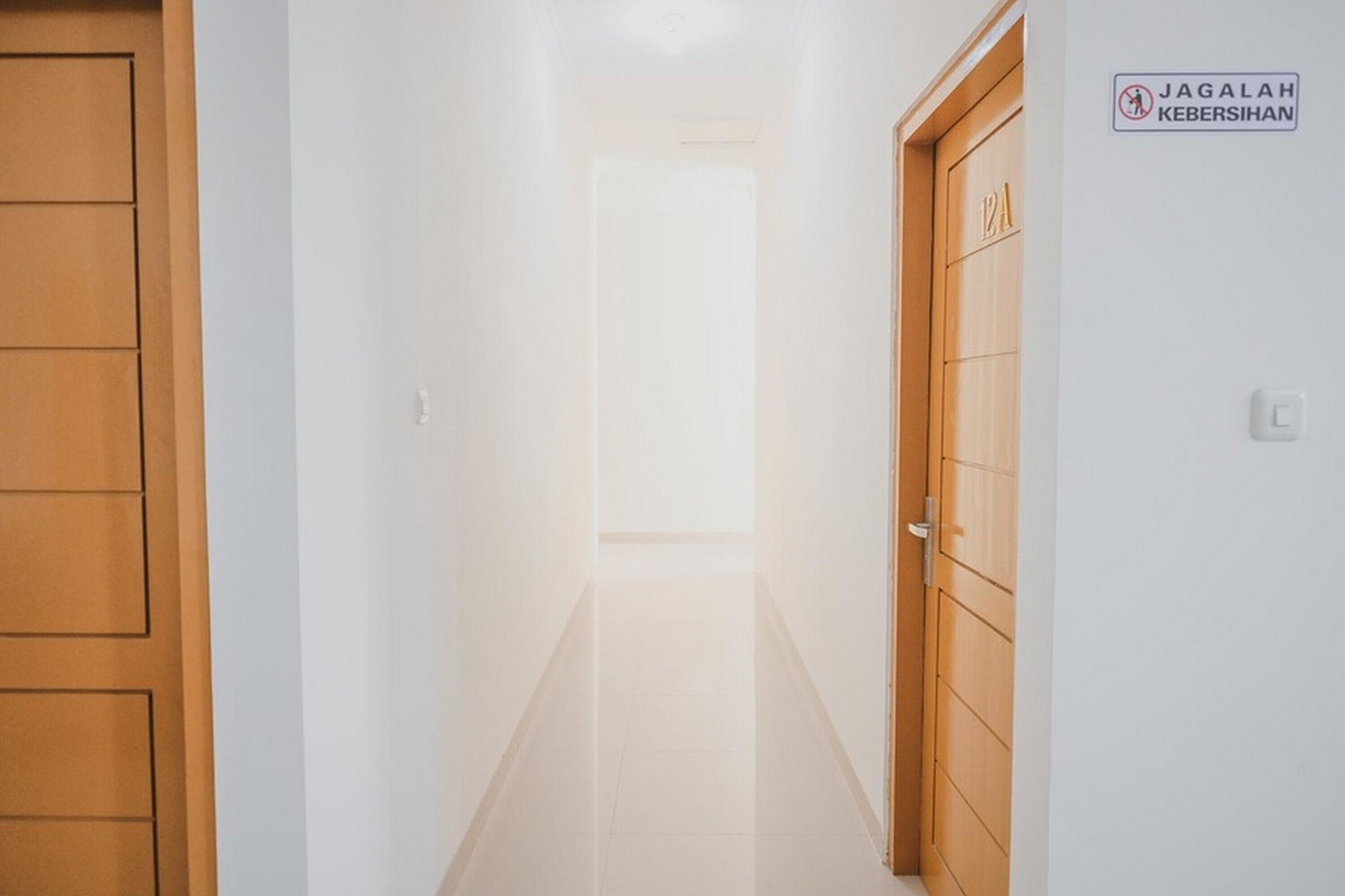 Un couloir avec une porte en bois sur la gauche et une pancarte indiquant « JAGALAH KEBERSIHAN » sur la droite.