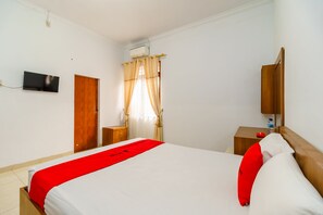 A hotel room with a bed, a wooden nightstand, a TV mounted on the wall, a window with curtains, and an air conditioning unit. - RedDoorz Syariah @ Jalan Dr Mansyur Medan (Medan)