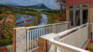 Un balcón con pared y barandilla de piedra, con vista a un mural de un paisaje con río y montañas.