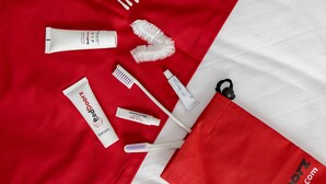 A red and white striped towel, a white tube of shampoo, a white tube of toothpaste, a white toothbrush, a white tube of cream, and a white hairbrush on a red fabric.