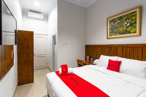 A hotel room with a wooden headboard, a bed with white bedding and a red accent, a painting on the wall, and a wooden wardrobe. - RedDoorz Near Universitas Muhammadiyah Yogyakarta (Kasihan)