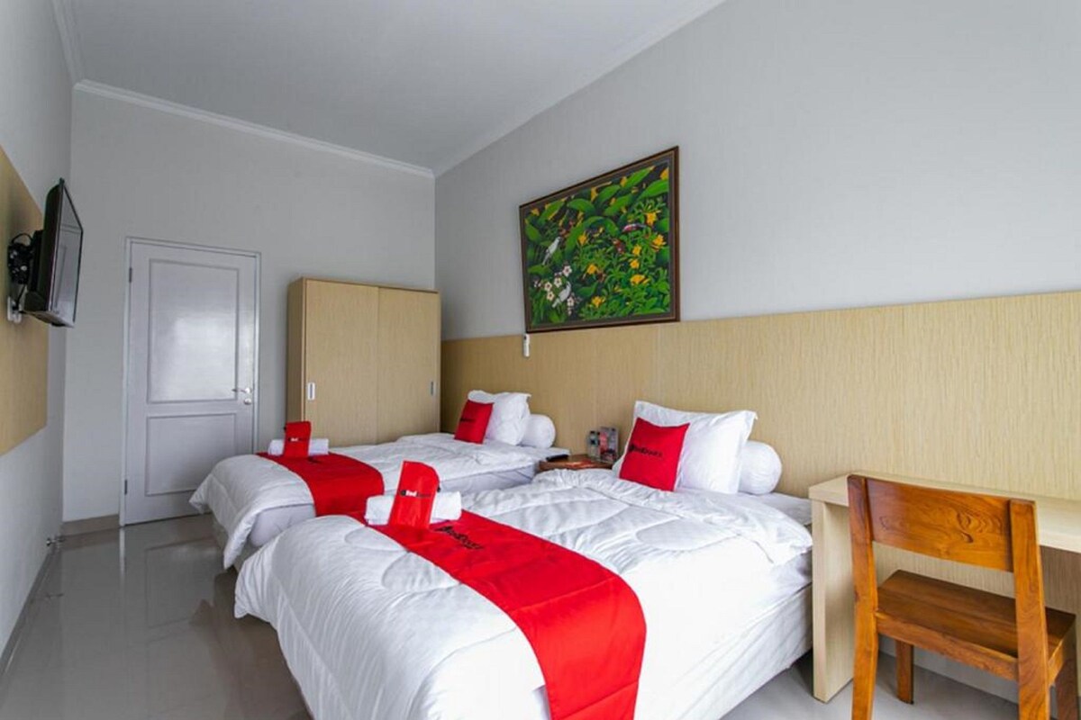 A hotel room with two beds, a wooden desk, a chair, a door, and a painting on the wall.