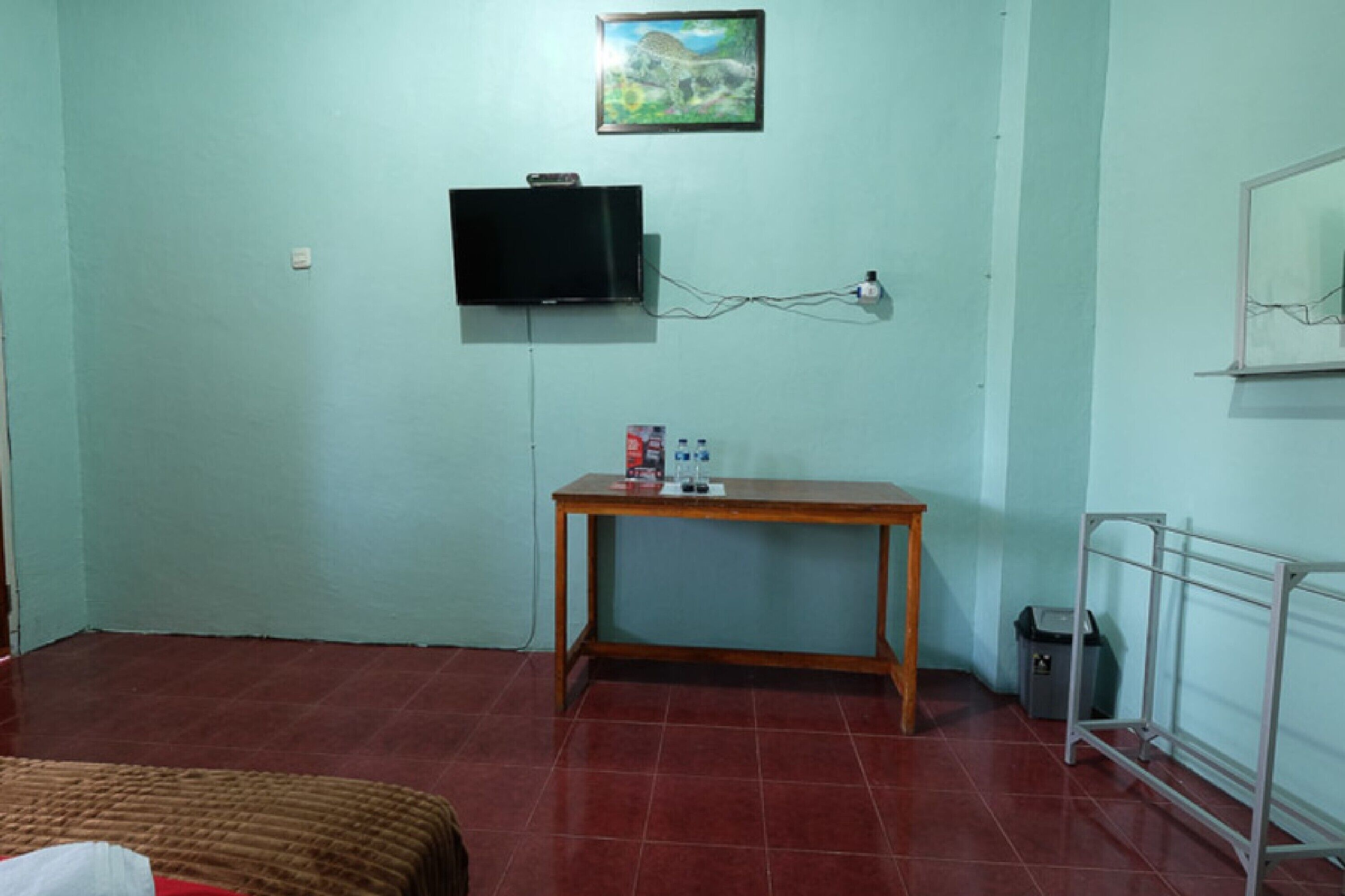 Una habitació amb un llit, una taula de fusta, una televisió penjada a la paret i un cubell de brossa.