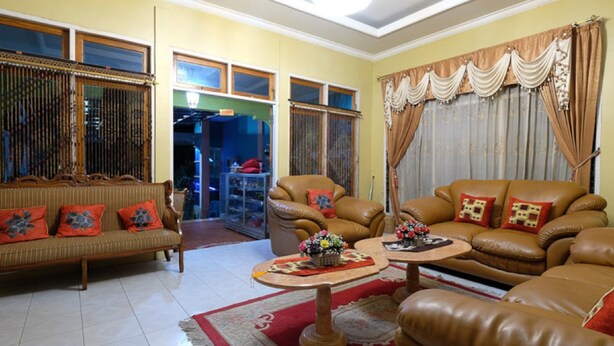 A living room with a sofa, armchairs, and a coffee table. There is a window with curtains and a door leading to another room.