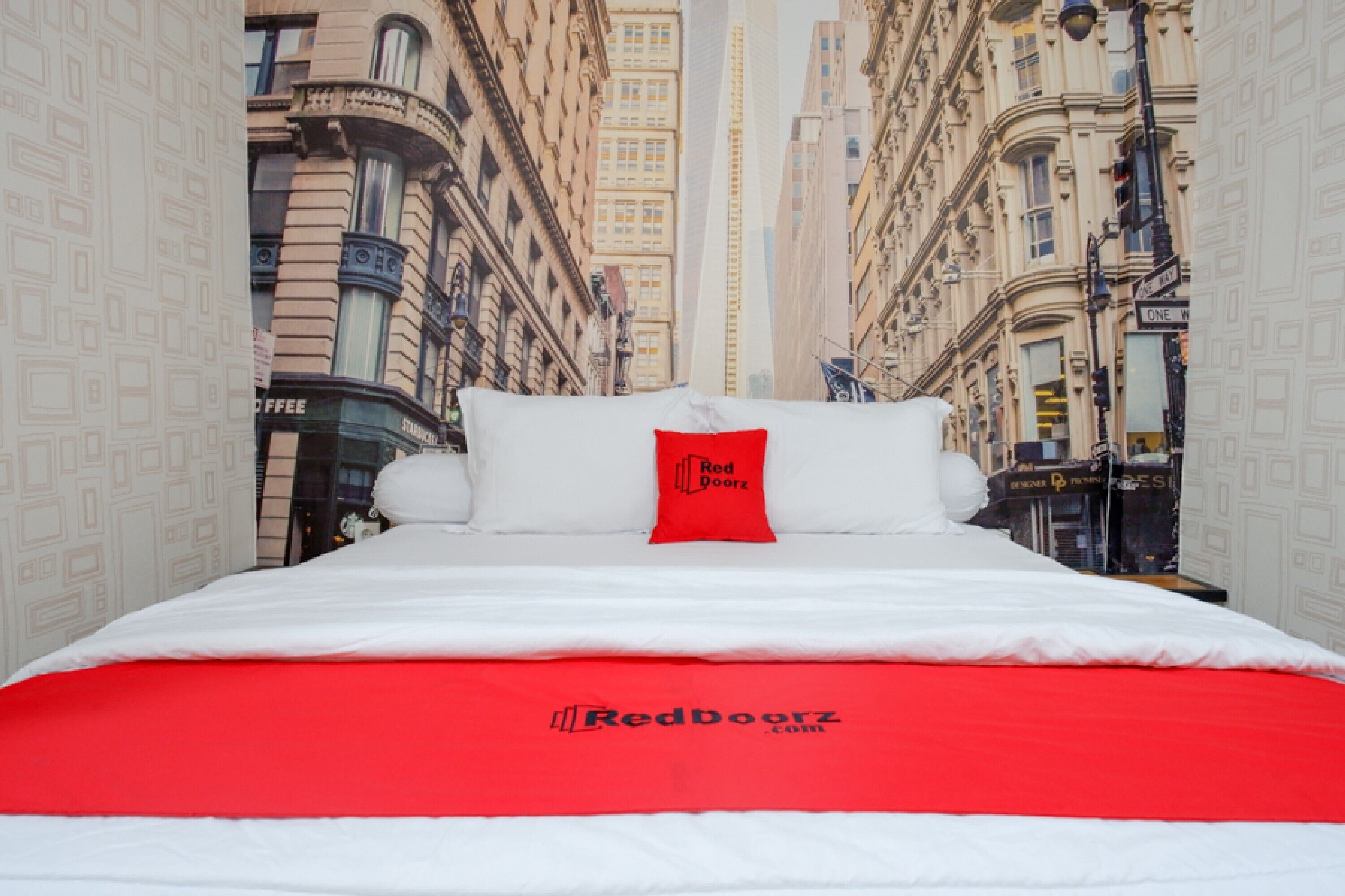 A bed with a red and white bedspread, a white pillow with a red logo, and a cityscape wallpaper in the background.