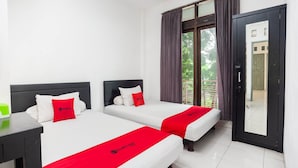 Quarto de hotel com duas camas, roupa de cama branca, detalhes em vermelho, uma mesa de cabeceira, uma janela com cortinas e uma porta que dá para área externa.
