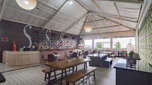Un restaurant spacieux avec de hauts plafonds, une salle à manger centrale dotée de tables et de bancs en bois, un comptoir où est affiché le menu, et une vue sur l’extérieur grâce à de grandes fenêtres.