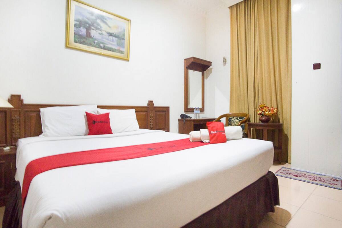 Quarto de hotel com cama bem arrumada, mesa de cabeceira com abajur, espelho e janela com cortinas.
