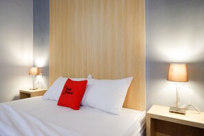 Ein ordentlich bezogenes Bett mit weißer Bettwäsche und einem roten Kopfkissen, an beiden Seiten flankiert von Nachttischen mit Lampen, in einem Zimmer mit Holz-Kopfteil und hellen Wänden.