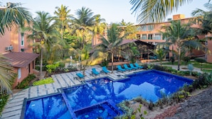A swimming pool with blue tiles, surrounded by lounge chairs and palm trees, with a building in the background. - Regenta Place Mandrem Beach-Resort (Mandrem)