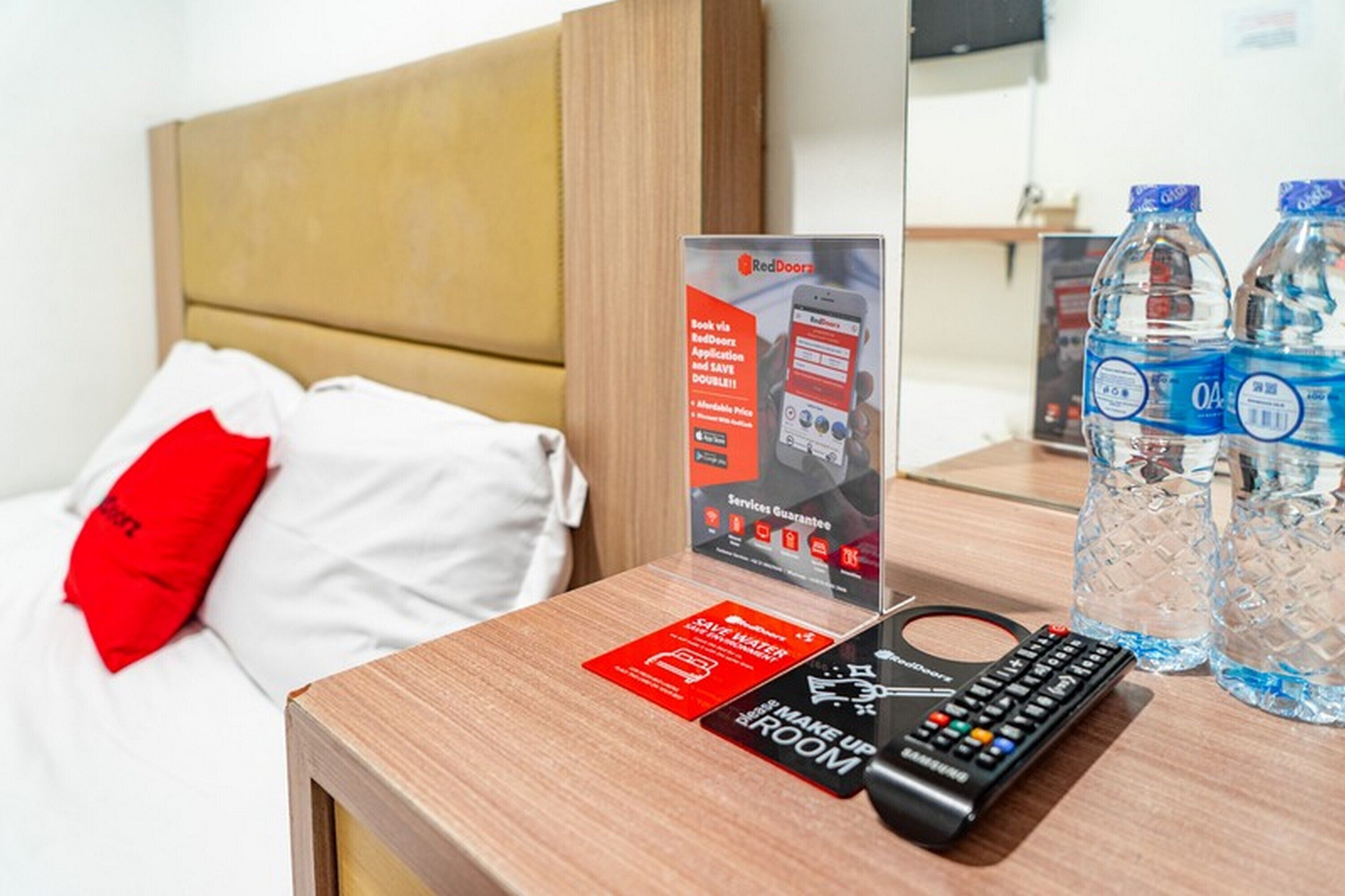 Quarto de hotel com uma cama, uma placa com os serviços RedDoor, um controle remoto e duas garrafas de água na mesa de cabeceira.