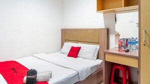 A single bed with a wooden headboard, a red pillow, and a white blanket. There is a desk with a mirror, a red stool, and a water bottle on it. - RedDoorz @ Kebon Kacang (Jakarta)