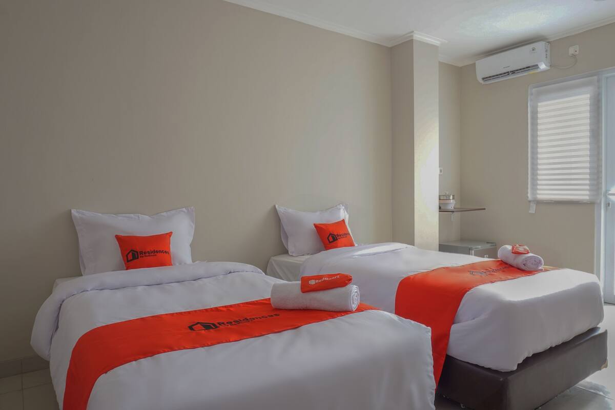 A hotel room with two beds, each with white bedding and orange accents, including pillows and a blanket. There is a window with blinds, an air conditioning unit on the wall, and a small table with a lamp.