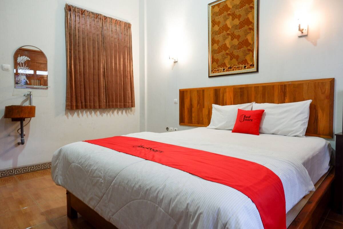 A hotel room with a wooden headboard, a large bed with white bedding and a red throw, a wooden side table, a mirror, and a patterned wall hanging.