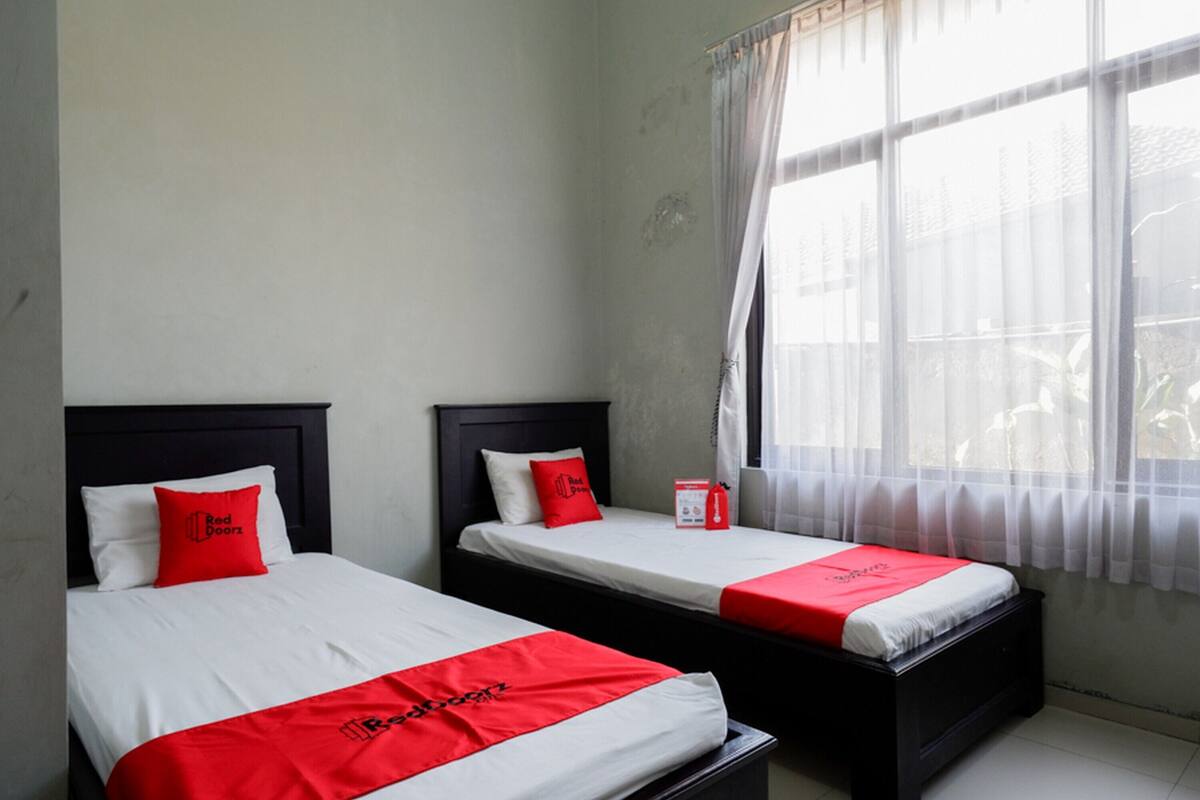 Two single beds with white and red bedding, a small bedside table, and a window with sheer curtains.
