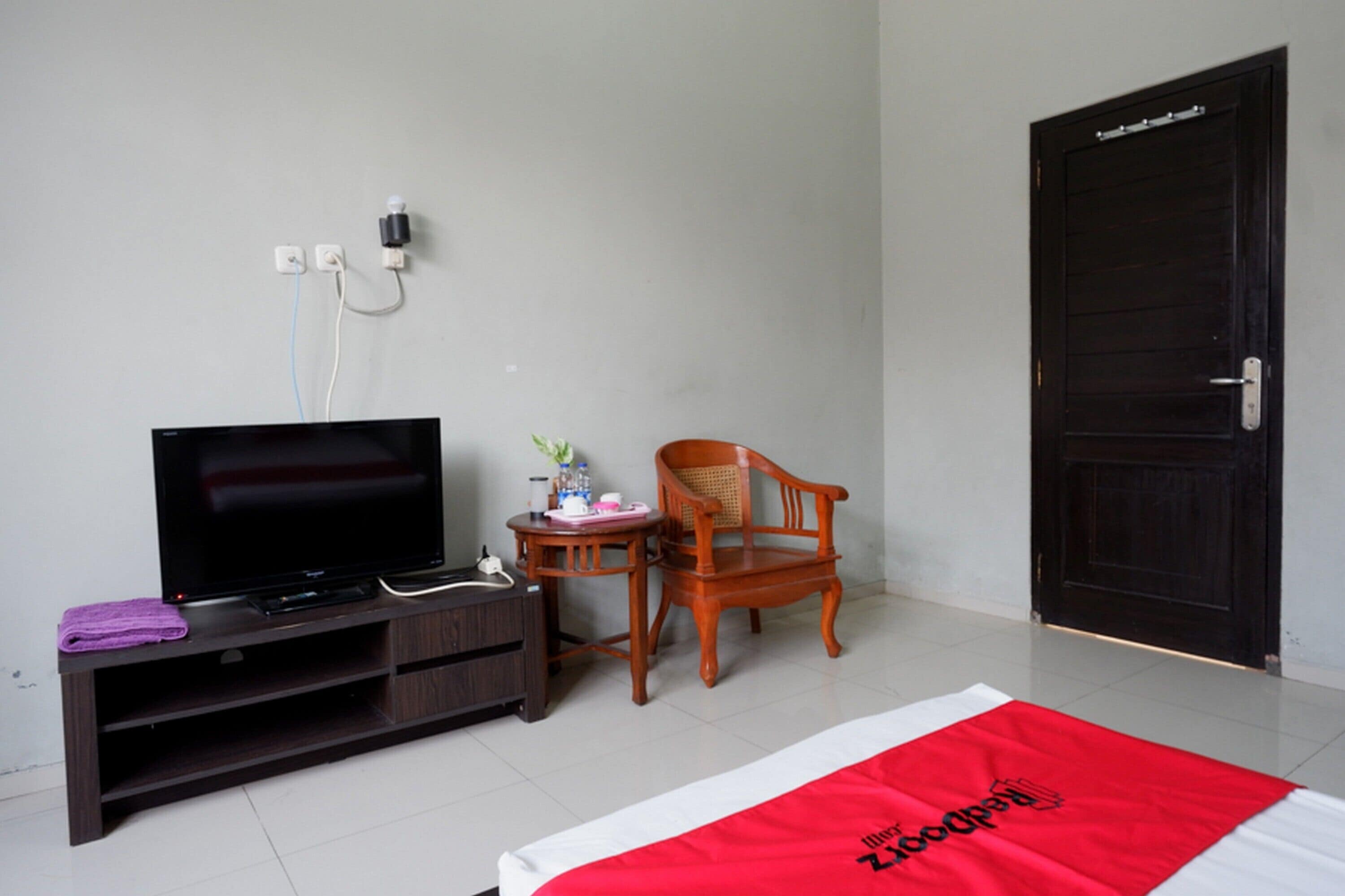 Ein Hotelzimmer mit Fernseher, einem Holzstuhl, einem kleinen Tisch mit Pflanze und einem Bett mit roter und weißer Decke.