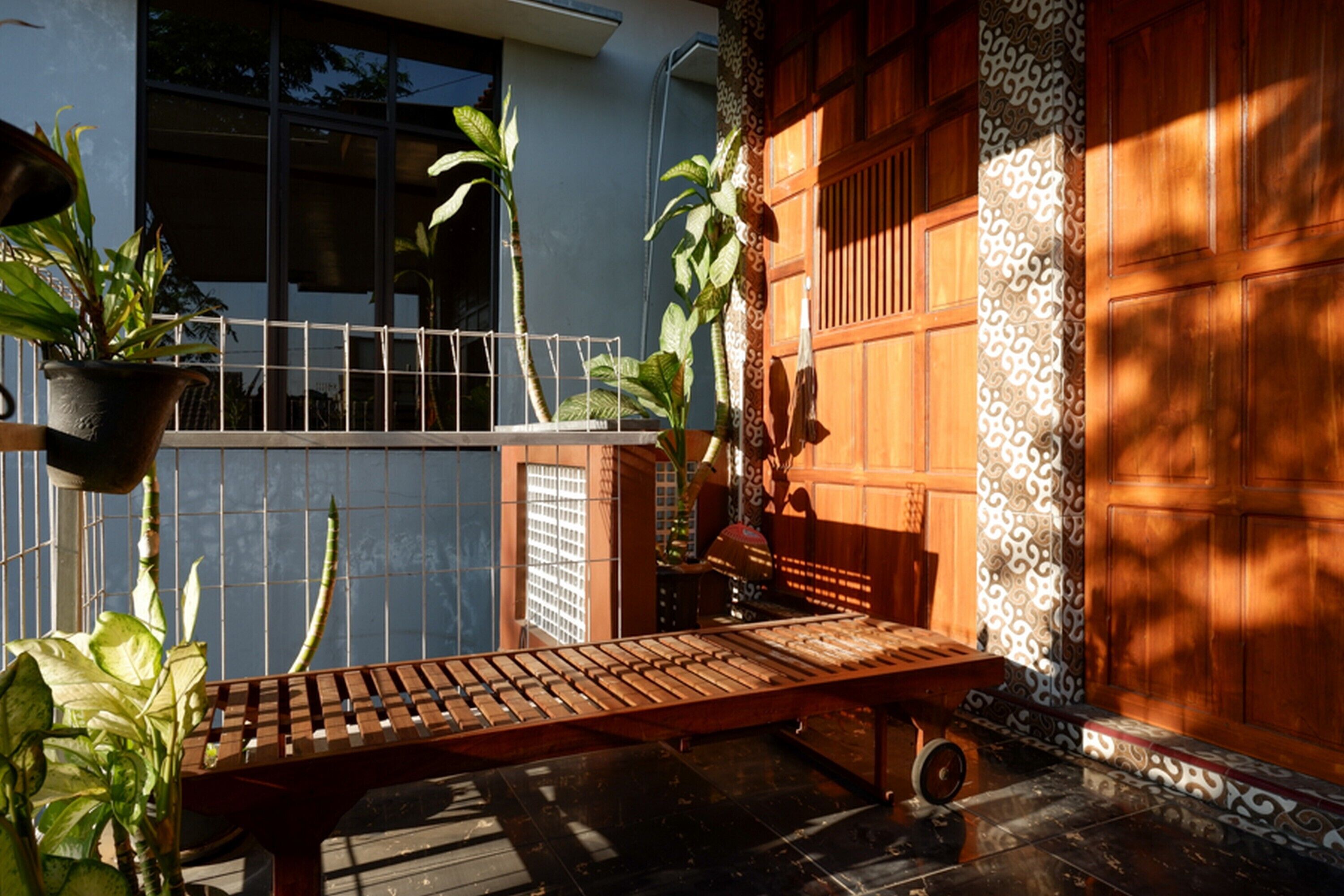 A wooden bench in a shaded outdoor area with potted plants and a patterned wall.