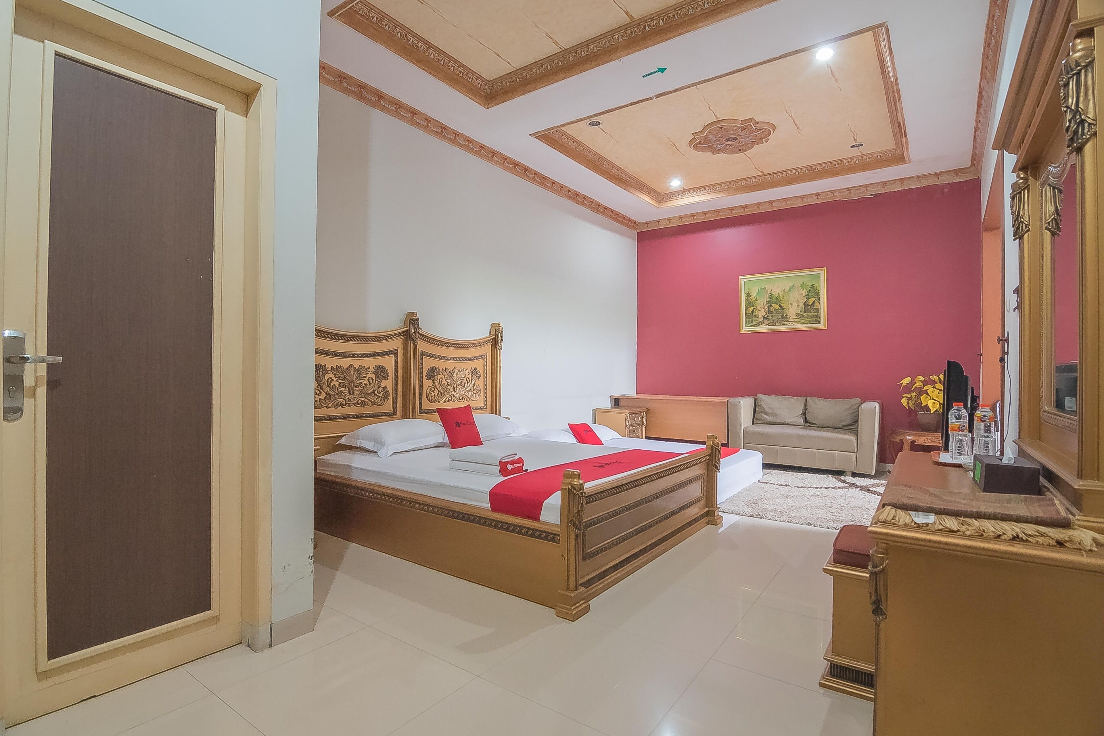 A bedroom with a wooden bed, a beige sofa, a red wall, and a ceiling with a decorative design.