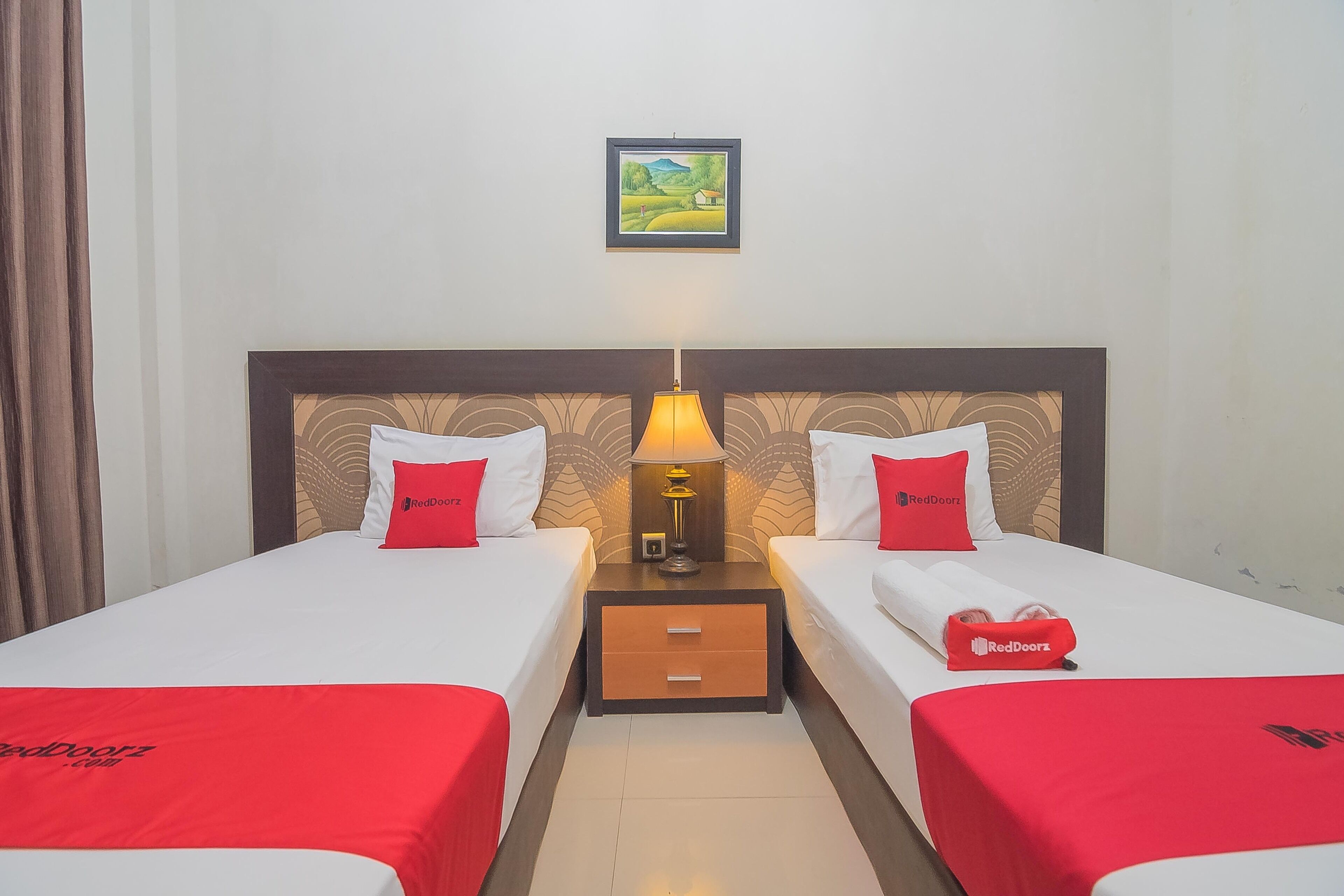 A hotel room with two beds, each with a red bedspread and matching pillows, a nightstand with a lamp, and a framed picture on the wall.