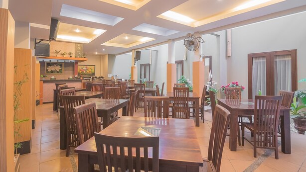 A spacious dining area with multiple wooden tables and chairs, recessed ceiling lights, and potted plants.