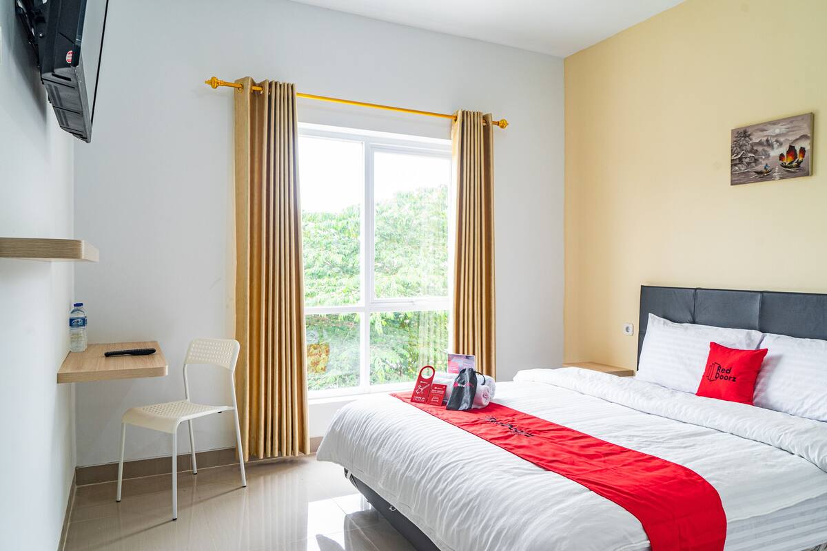 Ein Hotelzimmer mit Bett, Sessel, Schreibtisch, einem Fenster mit Vorhängen und einem Bild an der Wand.