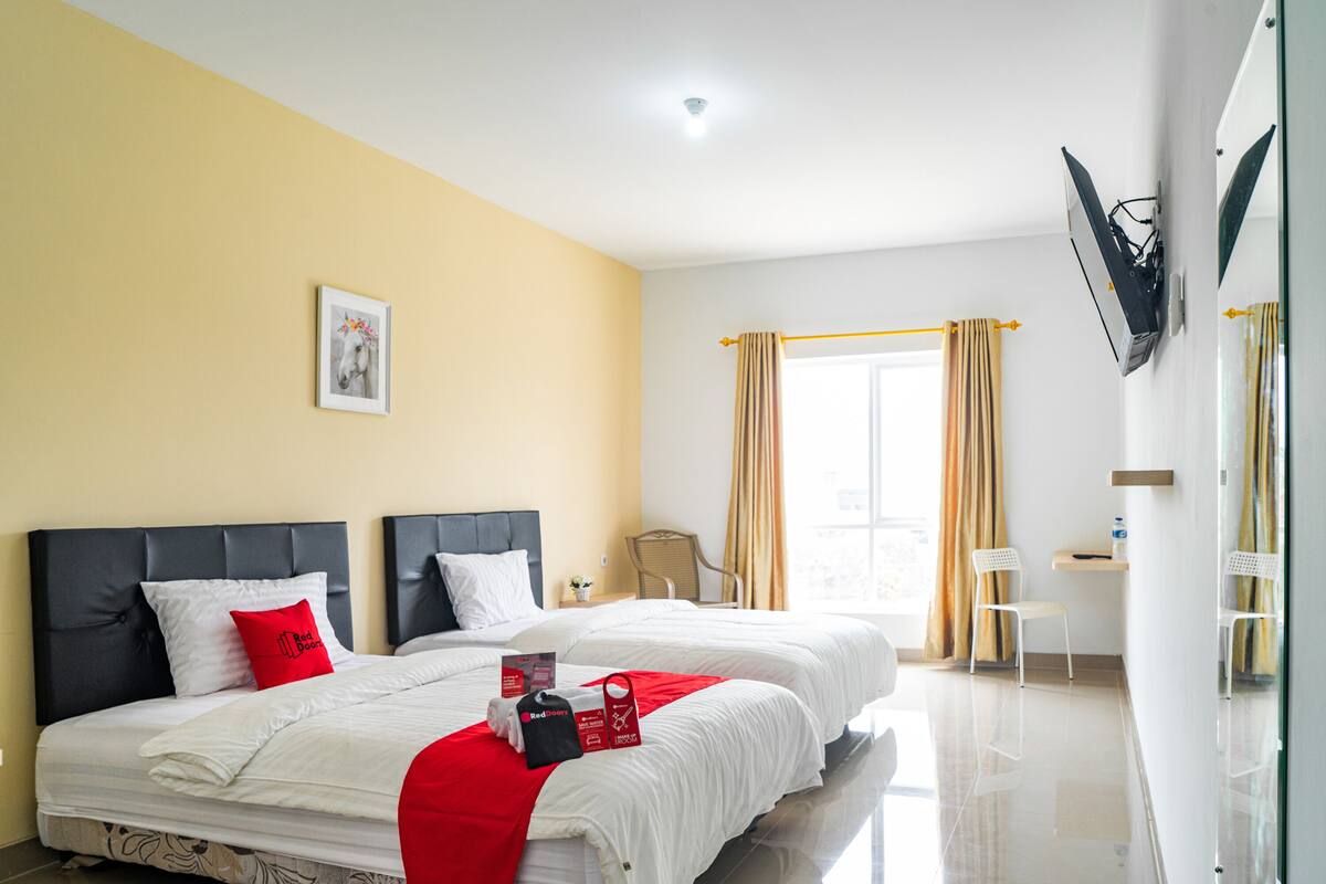 Ein Hotelzimmer mit zwei Betten, einem wandmontierten Flachbildfernseher, einem Fenster mit Vorhängen und einem kleinen Tisch mit einem Stuhl.