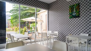 Un espace repas avec des tables et des chaises blanches, de grandes fenêtres donnant sur un paysage verdoyant et un bâtiment à l'extérieur, et un mur orné de motifs.