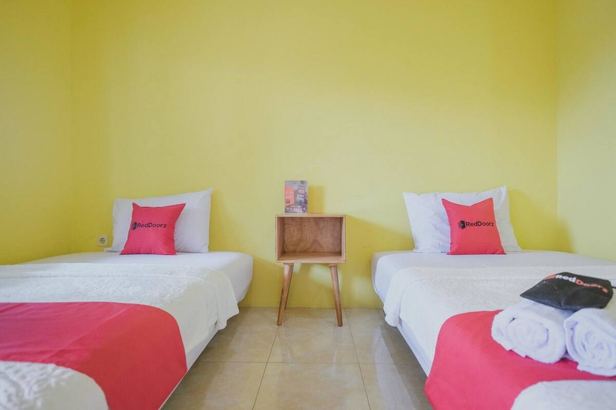 A hotel room with two beds, each with white bedding and red pillows featuring the RedDoorz logo. A small wooden nightstand with a box on top is positioned between the beds.