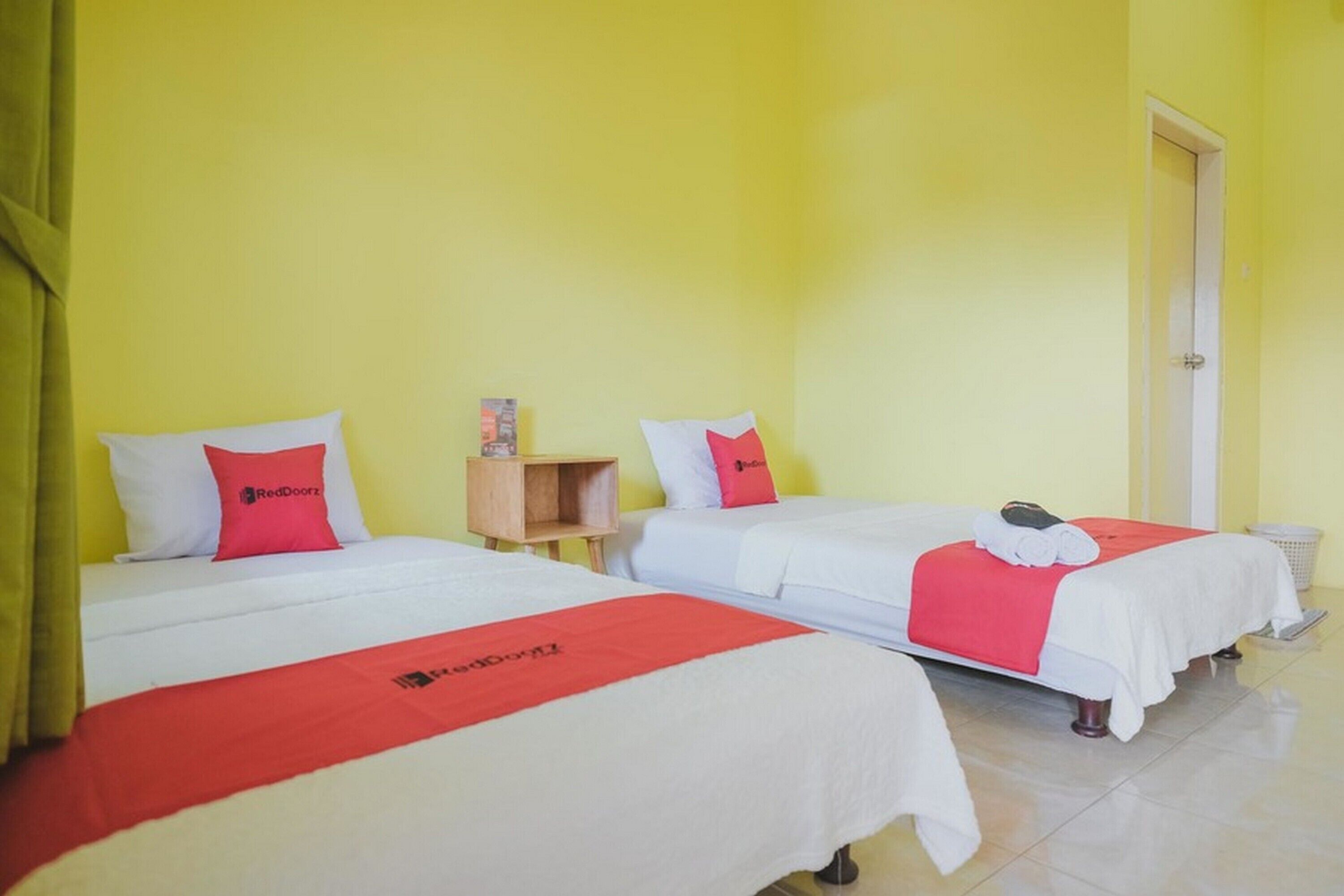 A hotel room with two beds, each with white bedding and red accents, a small wooden nightstand, and a door to the right.