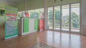 A reception area with a green and white counter, a poster on the left, and a view of trees through the glass doors. - RedDoorz Syariah near Museum Wisma Karya Subang (Subang)