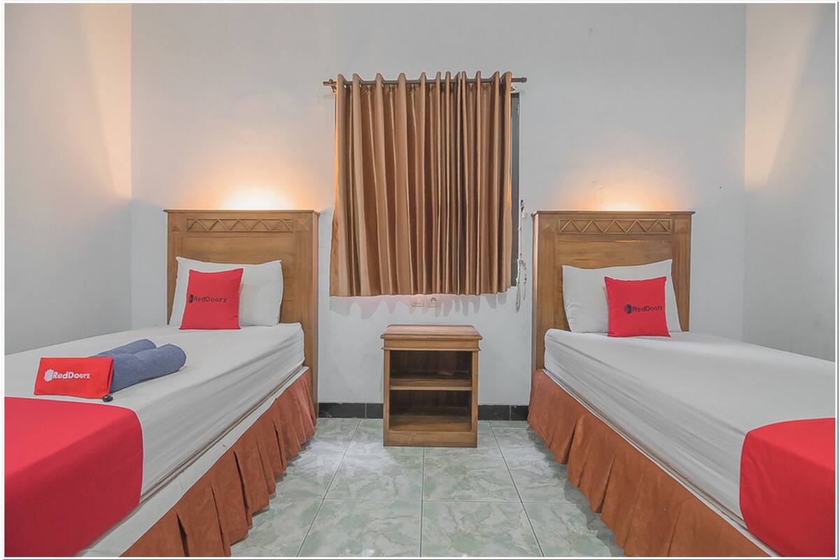 A hotel room with two single beds, each with a wooden headboard, a small wooden nightstand, and red bedding.