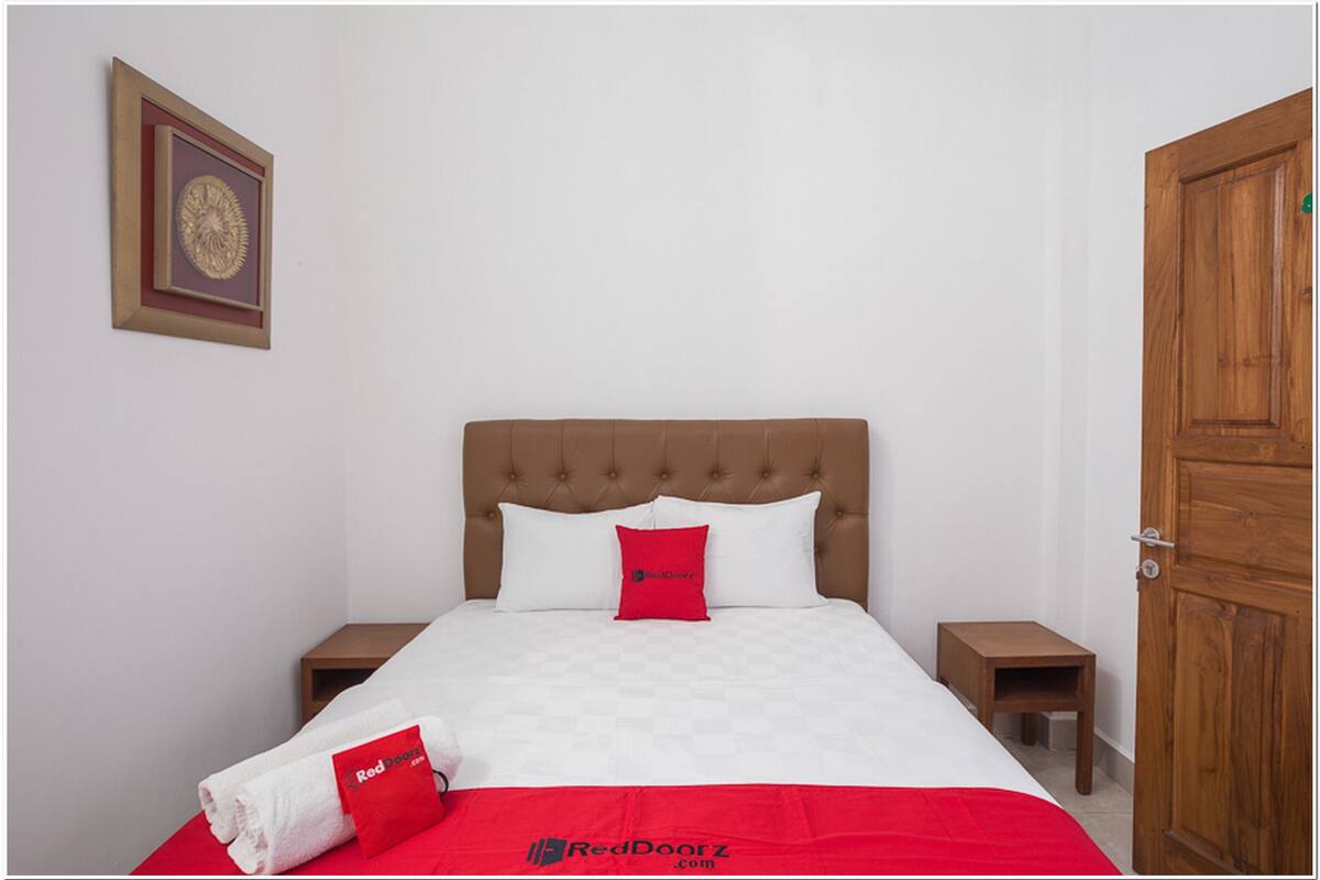 Een net opgemaakt bed met een rode sierkussen, een rode deken en een wit kussen. Aan beide zijden van het bed staat een klein nachtkastje, en boven het bed hangt een lijst met een foto.