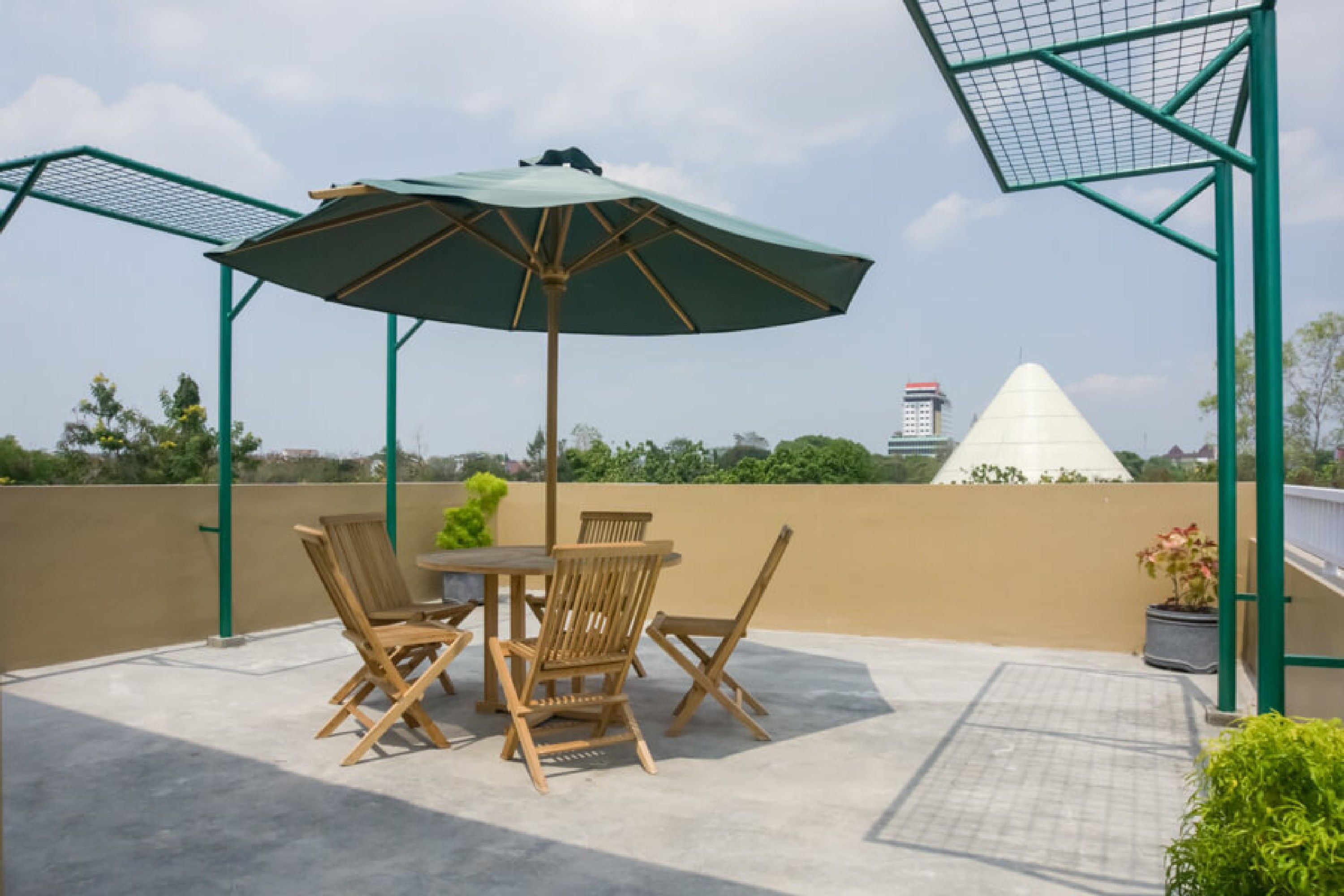 Um pátio externo com mesa e cadeiras, um guarda-sol verde e vista para um edifício em formato de pirâmide.