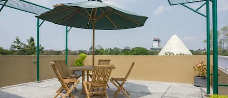 Um pátio externo com mesa e cadeiras, um guarda-sol verde e vista para um edifício em formato de pirâmide.
