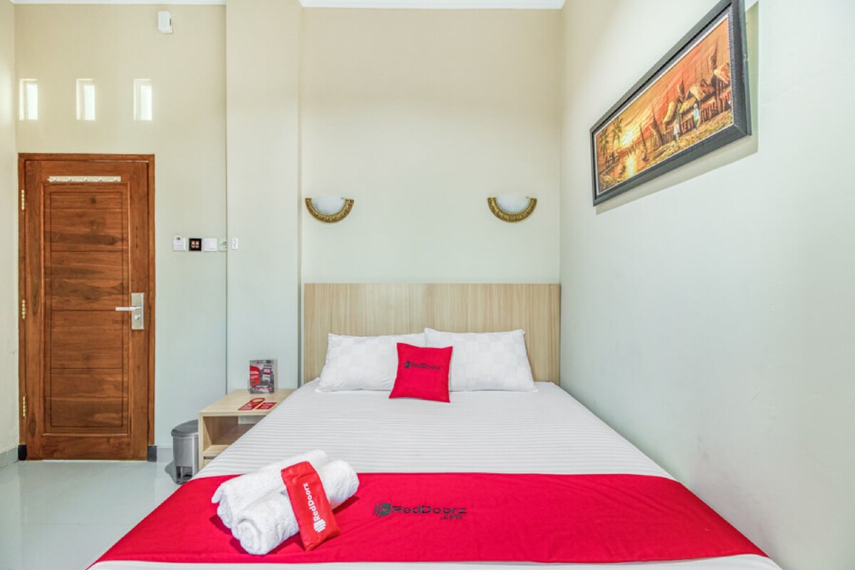 A hotel room with a bed, a wooden door, a nightstand, a framed picture on the wall, and a red blanket with a towel on the bed.