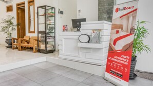 Ein Empfangsbereich mit einem weißen Empfangstresen, einem Holzstuhl, einer Glastheke und einem RedDoorz Werbeplakat.