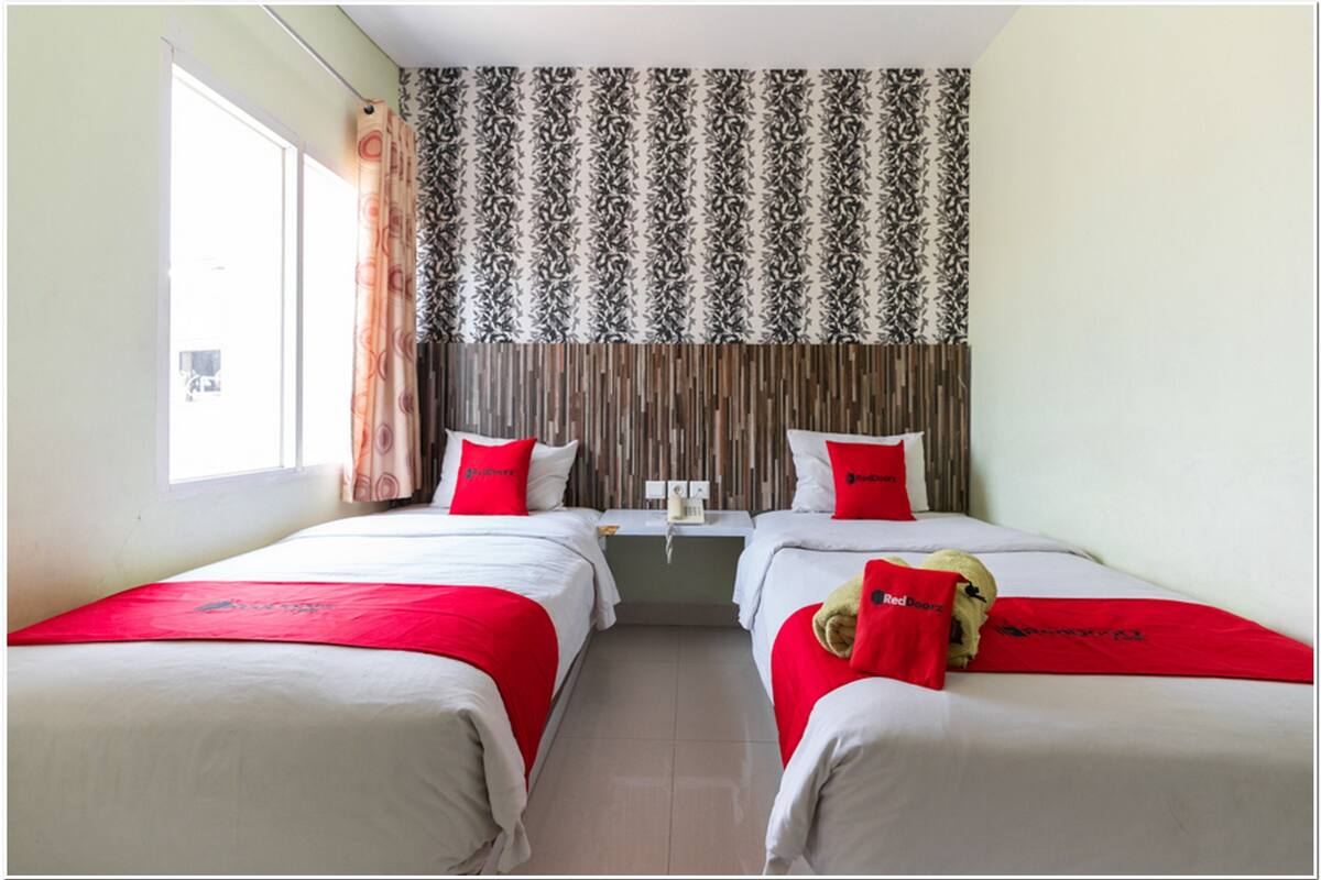 A hotel room with two beds, each with red bedding and pillows, a window with curtains, and a patterned wallpaper above the beds.