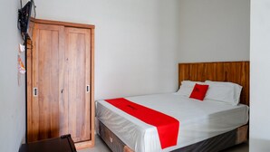 A bedroom with a wooden wardrobe, a bed with white bedding and a red accent pillow, and a black desk.