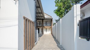 A narrow, paved pathway flanked by white walls and wooden fences, leading to a building with multiple balconies and a sloped roof.