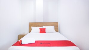 Een net opgemaakt bed met witte lakens en een rode deken, een houten hoofdbord en een klein nachtkastje met een rood voorwerp erop.