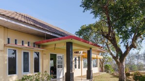 Un bâtiment jaune avec un toit recouvert de tuiles, un porche abrité avec des piliers noirs et un arbre en arrière-plan.
