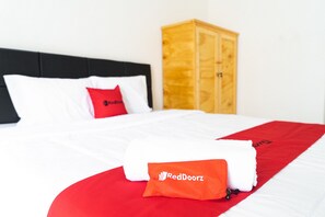 A neatly made bed with white and red bedding, a RedDoorz towel, and a wooden wardrobe in a hotel room.