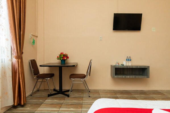 Habitación de hotel con cama, televisor montado en la pared, una pequeña mesa con sillas, una planta en maceta y una estantería con botellas.
