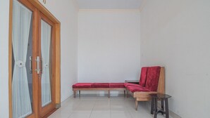 Quarto pequeno e limpo, com porta de madeira, um banco com almofada vermelha, uma mesa pequena e uma cadeira.