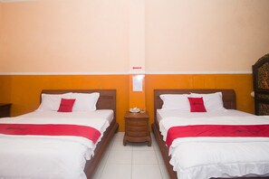 Habitación de hotel con dos camas, cada una con ropa de cama blanca y almohadas rojas, una mesita de noche de madera entre ellas, y una pequeña mesa con una lámpara.