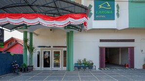 Ein Hotel Eingangsbereich mit überdachtem Bereich, einem Schild für 'Larona Hotel' und einem Parkplatz mit Motorrad und Pflanzen in Töpfen.