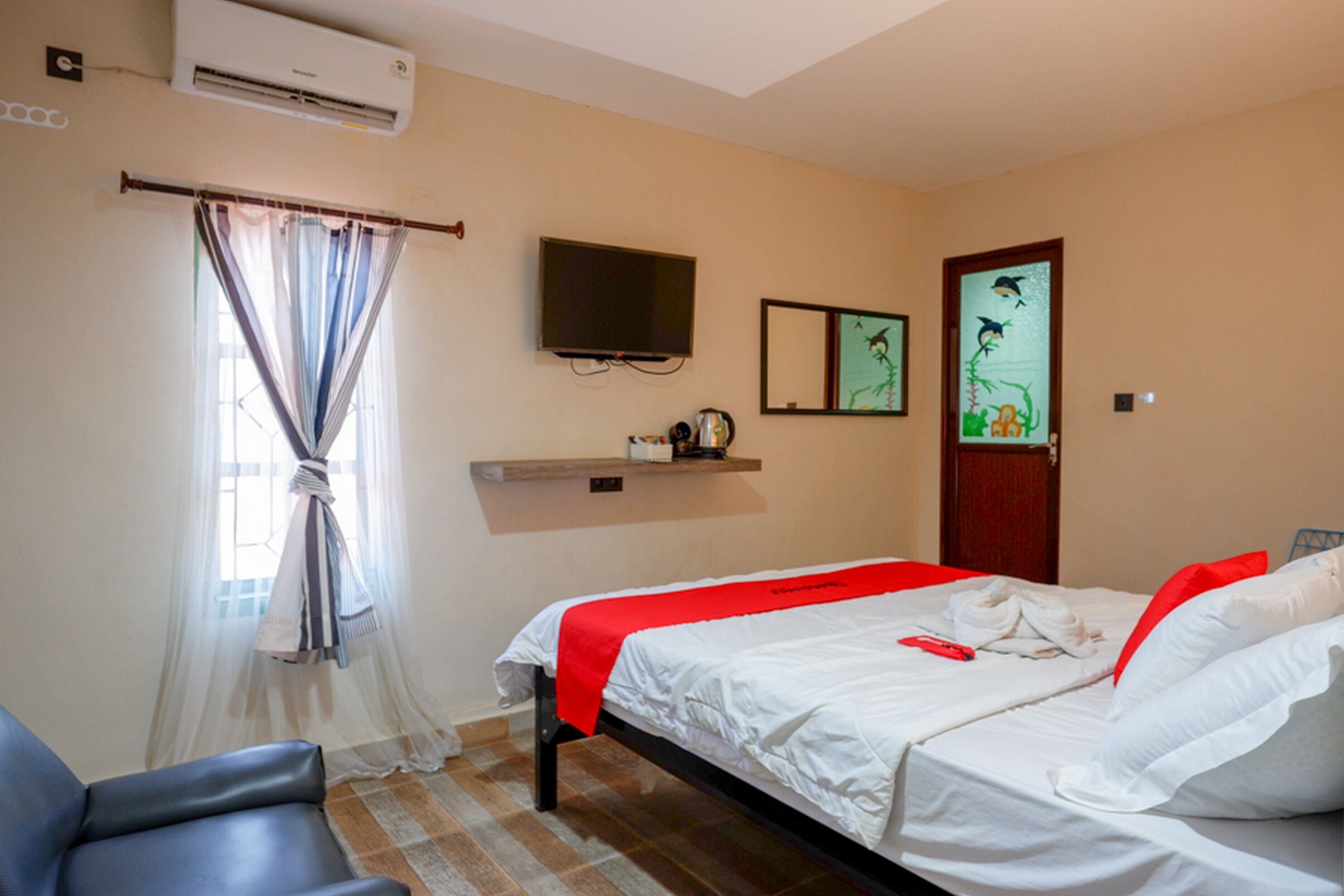 A hotel room with a bed, a chair, an air conditioner, a television, and artwork on the wall.