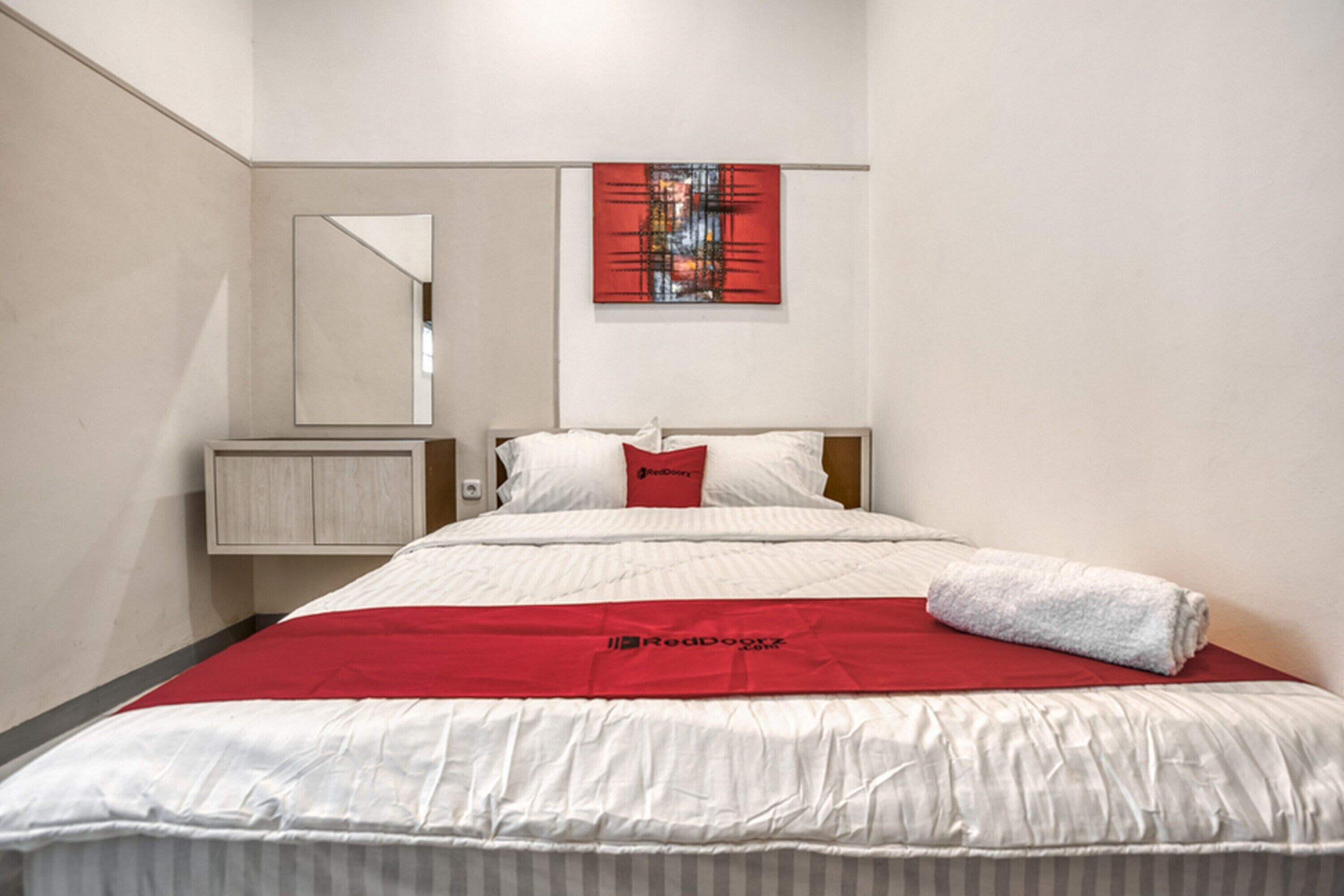 A hotel room with a bed, a red throw blanket, a red pillow, a mirror, and a red artwork on the wall.