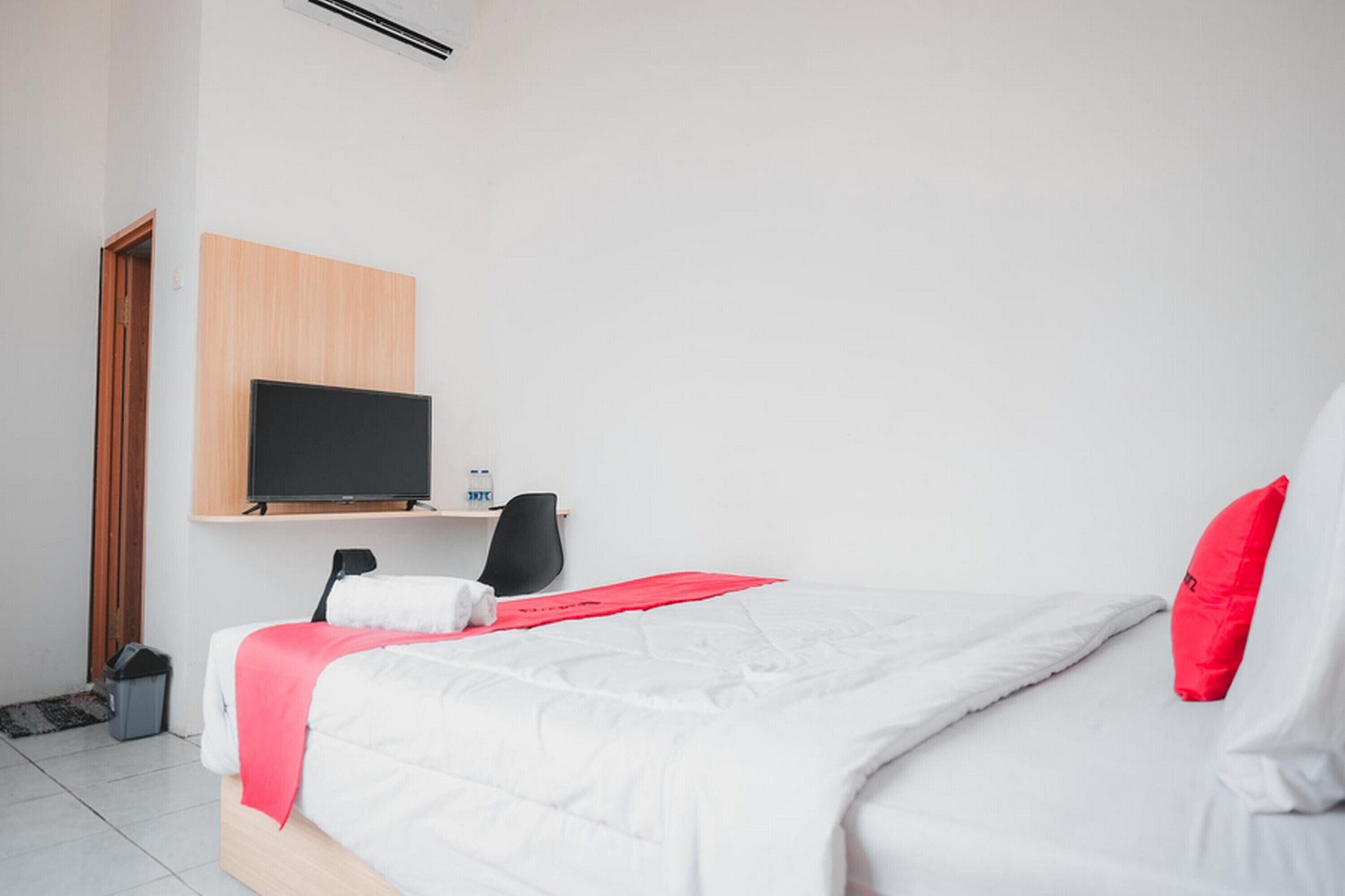 A hotel room with a bed, a TV mounted on the wall, a desk with a chair, and a red pillow.